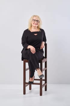Middle age woman in black outfit and glasses on chair, studio shot