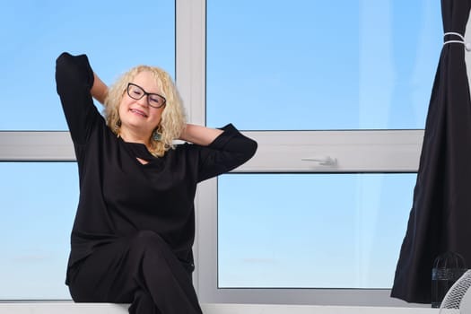 Happy middle age woman in glasses sits on windowsill