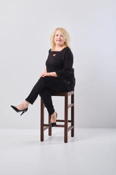 Middle age woman in black outfit sits on chair in white studio