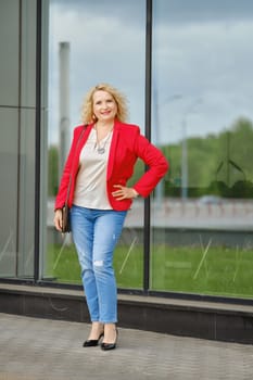 Confident woman in red jacket, blouse and denim jeans standing near the reinforced glass wall of the building