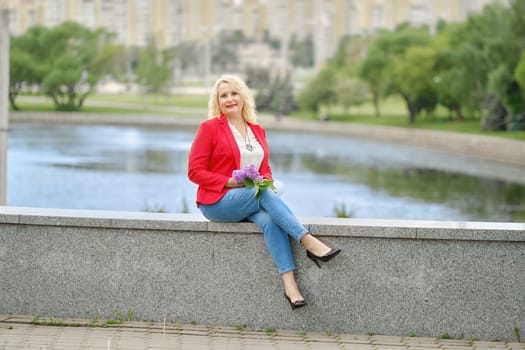Positive middle-aged woman is sitting on the embankment in a city park