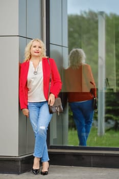 Confident woman in red jacket, blouse and denim jeans stands near the reinforced glass wall of the building
