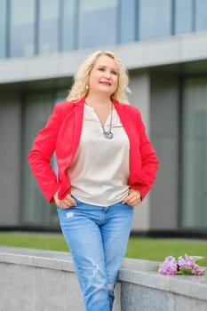 Confident senior woman in red jacket, blouse and denim jeans standing near the modern building