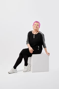 Happy old woman with short pink hair in long knitted dress sits on white box in the studio