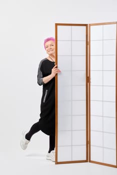Playful old woman with short pink hair looks out from behind a screen in white room