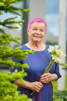 Portrait of smiling mature woman with short pink hair standing near fir-tree with spring flowers in hands outdoors