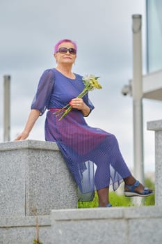Outdoor portrait of smiling mature woman with short pink hair with spring flowers in hands