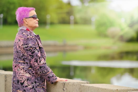 Funny aged woman with short pink hair and woolen coat standing by the river embankment in city park