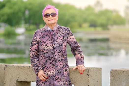 Active aged woman with cheerful smile standing by the river embankment in city park