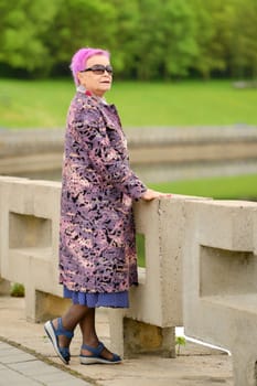 Full length portrait of old woman with short pink hair and woolen coat standing by the river embankment in city park