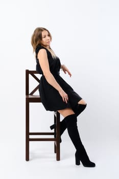 Stylish confident middle aged woman wearing black dress and suede boots sitting in profile on tall chair in studio