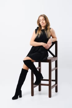 Fashionable trendy senior woman in loose black dress and suede boots sitting on tall chair in studio over white background