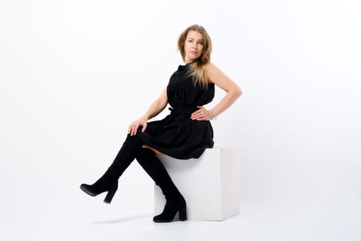 Fashionable elegant senior woman posing in studio sitting on white cube in profile
