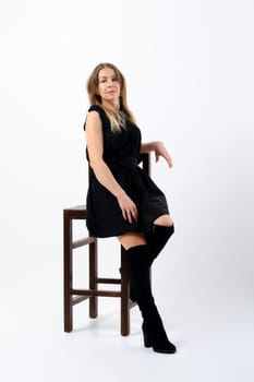 Stylish confident senior woman wearing black dress and suede boots sitting on tall chair in studio