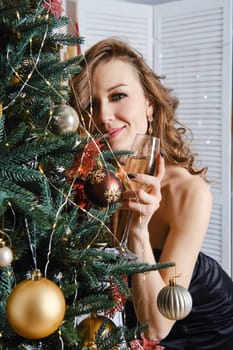 Beautiful young woman leaning to the christmas tree with a glass of champagne in hand