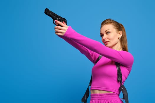 Stylish woman in purple knitted pantsuit aims a gun over blue studio background. Fashion police and accurate hit on target concept.
