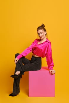 Playful woman in bright old-fashioned clothes sits in profile on purple cube over bright yellow studio background