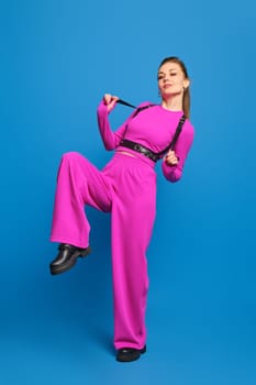 Joyful woman in purple knitted pantsuit pulls the belts of leather shoulder harness over blue studio background