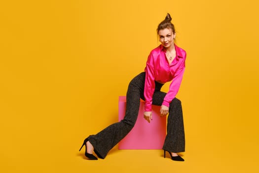 Tricky woman in bright disco-style clothes sitting on purple cube in studio on yellow background. Humorous and funny concept of playful mood.