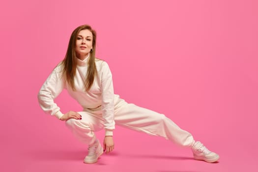 Active woman in white sportswear sits on squats and leaning on one leg over pink studio background