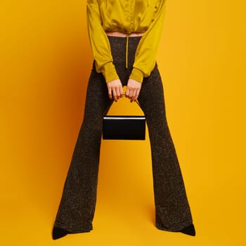 Female legs in flared trousers stand wide apart and hands hold a purse between them on yellow studio background