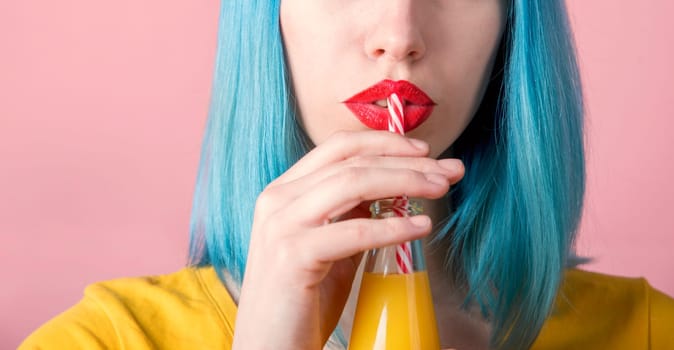 Closeup studio portrait of hipster young woman with red lipstick and blue dyed hair drinking orange juice from retro bottle with straw. Closeup of female mouth drinking summer cocktail. Trendy pastel color background.