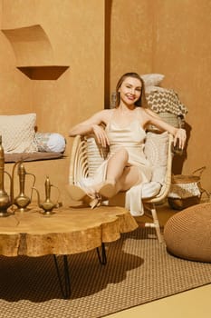 Smiling young woman sitting in wicker armchair with her legs on tea table in an oriental style interior and holding cup in hand