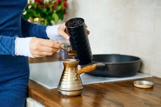 Closeup view of female hand with spoon with ground coffee near a turk. Making coffee in metal pot.