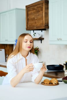 Pensive young woman sits behind the kitchen table with cup of coffee and thinking about something