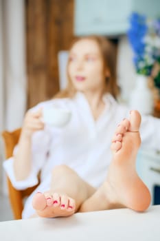 Closeup view of female feet on kitchen table (blurred woman holds cup of coffee on background)