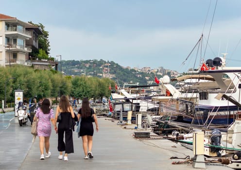 Istanbul, Turkey - August 31, 2022: Pedestrians walking on side walk by the Bosphorus Strait between Bebek and Hisari districts, beside docked motor boats and yachts, against blue sky on sunny day