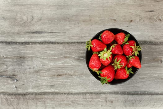 Tabletop view, small black ceramic bowl with strawberries, gray wood desk under. Space for text on left side.