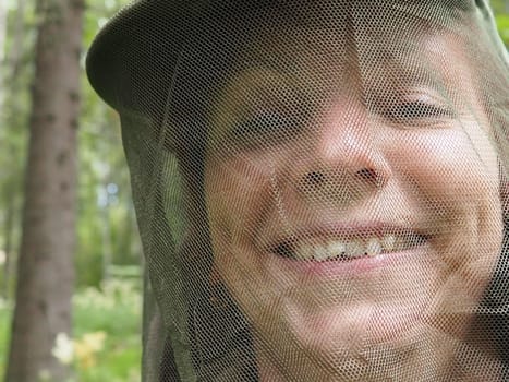 A woman in her 40s wearing a mosquito headdress smiles and looks at the camera. Cap and mesh against insects, mosquitoes, bees, wasps. Hunting and fishing in the spruce forest.