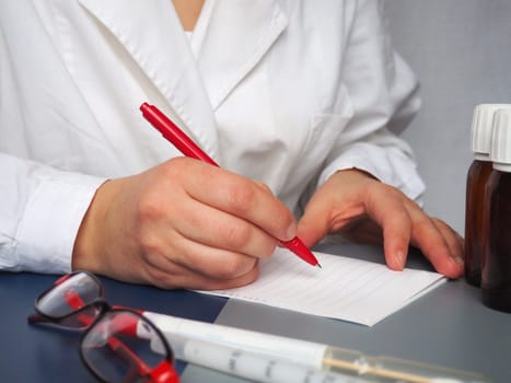 doctor writes down the prescription on paper. Medicines, glasses, a test tube, a syringe lie on the desktop. Pen in hand. A white coat is worn by a medical worker. Hands of a doctor or nurse.