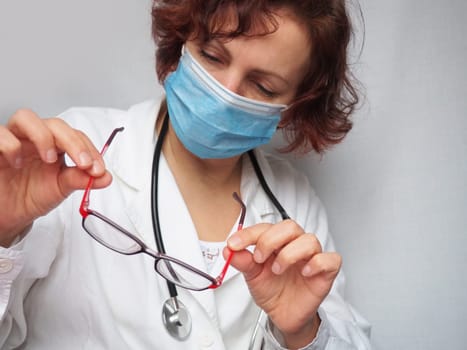 Doctor with stethoscope examining red-rimmed glasses. Medicine and healthcare. A frowning, concentrated face in a protective mask. An ophthalmologist or cardiologist. Gray background.