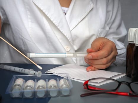 Hands of a doctor or nurse. Virus and bacteria test in hand. A white coat is worn by a medical professional. Medicines, glasses, test tube and a syringe are on the table. Medic at work on diagnostics