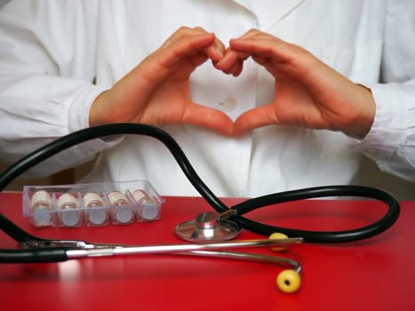 Doctor cardiologist with a stethoscope shows the heart with his hands. Stethoscope and medicines on the table. Hands gesture. Heart shape made by palms and fingers. Prevention of cardiac diseases.