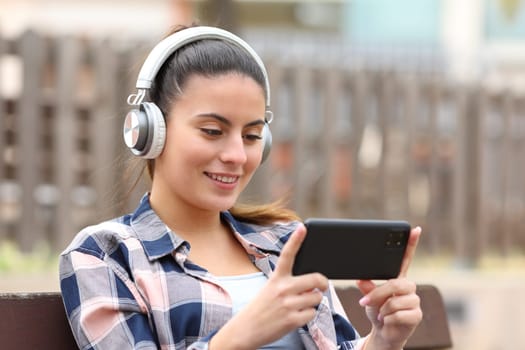 Happy teen wearing headphones watching media on phone in a bench