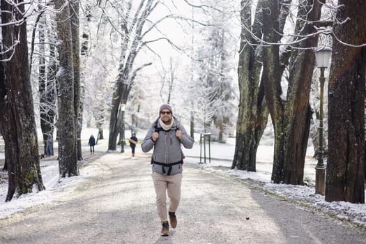 Sporty father carrying his infant son wearing winter jumpsuit and cap in backpack carrier walking in city park in winter.