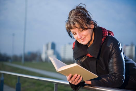 Side-body view of a mature woman with black leather jacket leaning on a railing in a book reading against a fuzzy urban background