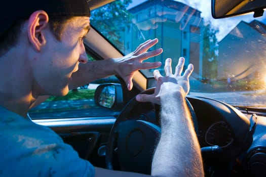 Side wide-angle upper body interior view of a blinded motorist (young man) holding his hands protectively in front of him towards the windshield with shocked look