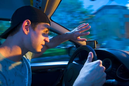 Side wide-angle upper body interior view of a blinded motorist (young man) holding his hands protectively in front of him towards the windshield with shocked look