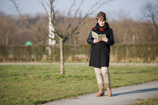 Side full-body view of a mature woman in black leather jacket standing on a path in the city park and reading a book