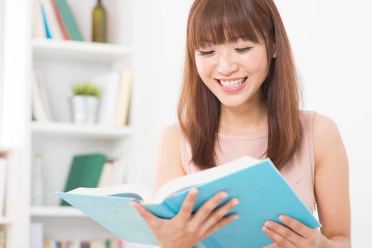 Portrait of happy Asian college girl laughing while reading on book. Young woman at school library.