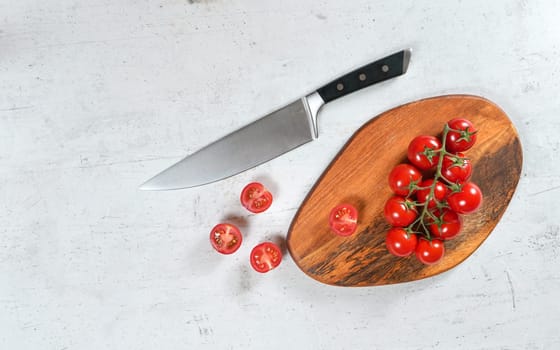 Vibrant small red tomatoes with green vines on wooden chopping board, large chef knife near, white stone table under, view from above.
