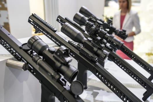 Black shooting scope optics mounted on metal bar displayed at weapons exhibition fair, closeup detail to adjustment knobs.