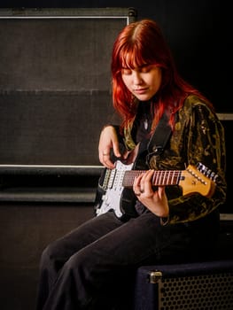 Young redhead woman playing electric guitar in front of large speaker cabinets.
