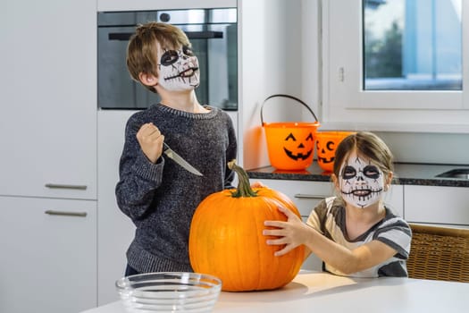 Two young children with skull face paint cutting a pumpkin in their kitchen.