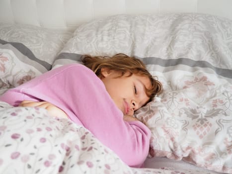 A beautiful caucasian girl of 8 years old with blond hair, dressed in pink pajamas, sleeps on a bed with a fluffy blanket, hugging a pillow. Soft light of the morning sun streams through the window