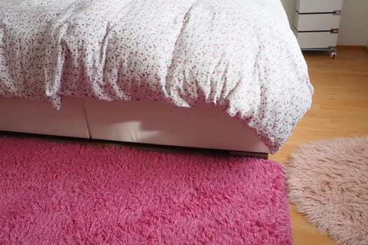 Cozy compact bedroom, white furniture, pink carpet and tulle curtains. Fluffy feather blanket thrown over the double bed. Romantic morning. Light from the window. The blanket hangs down to the floor